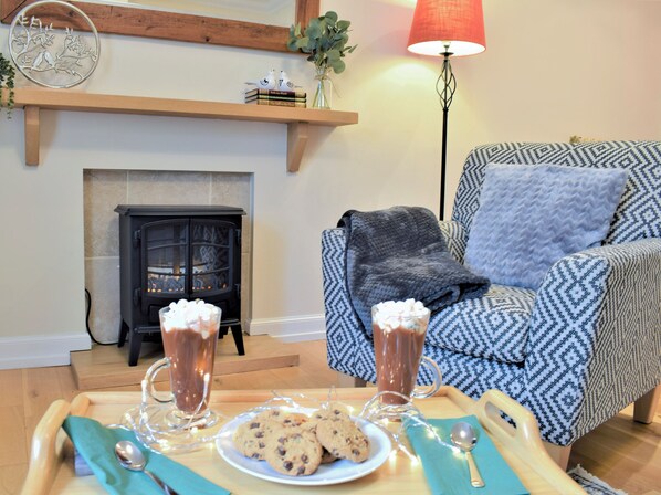 Un salon chaleureux avec une cheminée, un fauteuil à motifs, un plateau avec des biscuits et des boissons, et des œuvres d'art décoratives accrochées au mur.