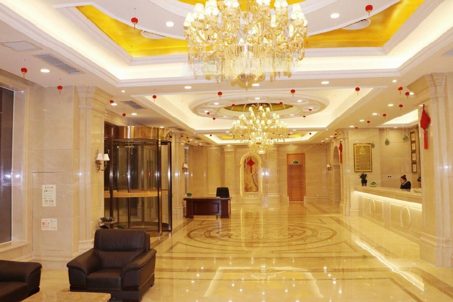 Un vestíbulo de hotel con recepción, un candelabro, un ascensor de cristal y una zona de estar.