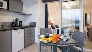 Une cuisine moderne équipée d’un lave-vaisselle, d’une machine à café et d’un four à micro-ondes. On y trouve un coin repas avec une table mise pour le petit-déjeuner, comprenant des fruits, une bouteille de jus et une tasse. On aperçoit un lit donnant sur le balcon à travers une porte vitrée.