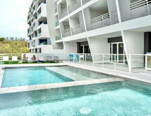 Un hotel moderno con piscina, mobiliario exterior y un edificio de varios pisos con balcones.