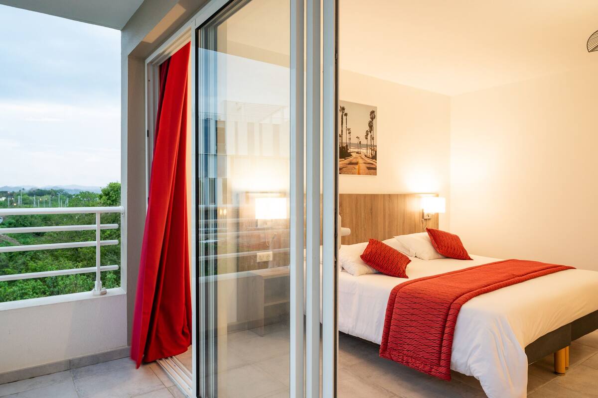 Habitación de hotel moderna con balcón, cama con ropa de cama blanca y roja, y vistas a zonas verdes.