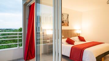 Habitación de hotel moderna con balcón, cama con ropa de cama blanca y roja, y vistas a zonas verdes.