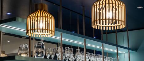 Un interior moderno con dos lámparas colgantes, un estante de cristal con varias copas de vino y un techo iluminado en azul.