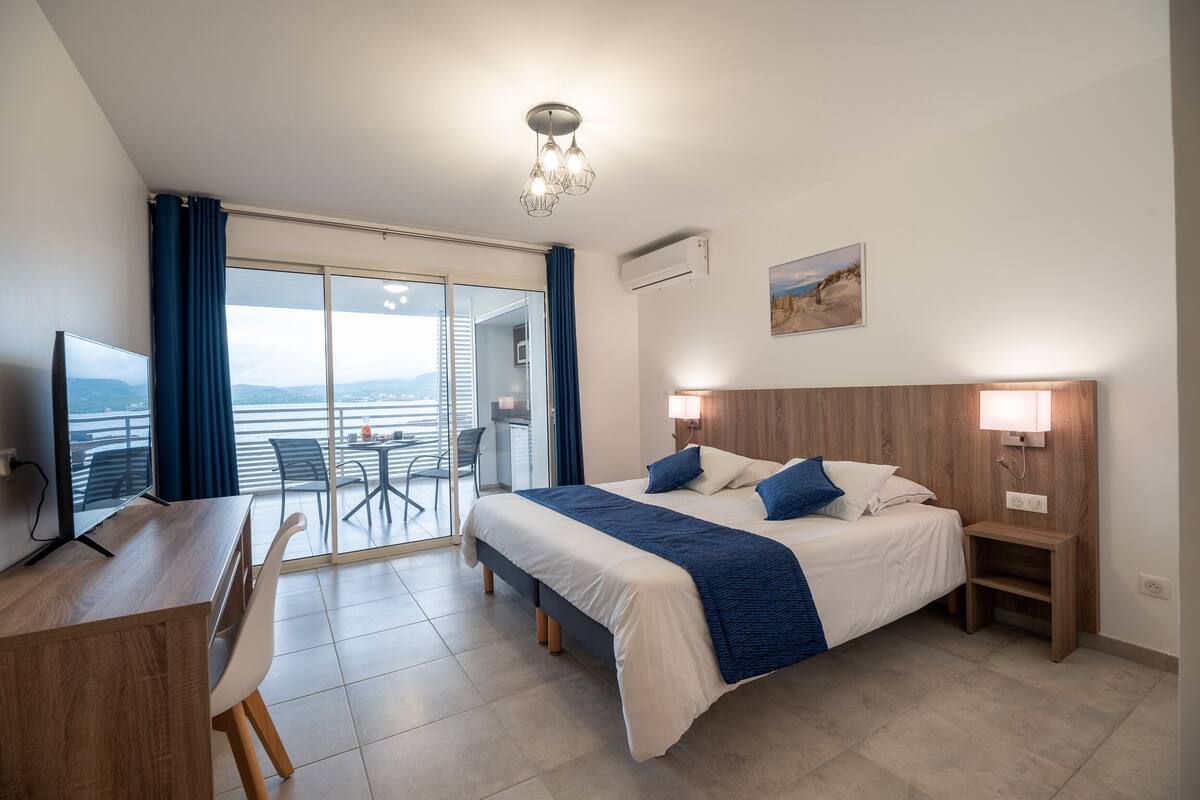 Habitación de hotel con cama, mesita de noche, escritorio y silla. Amplia ventana con vista a la ciudad y balcón con sillas.