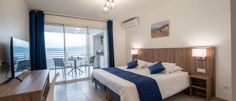 Une chambre d’hôtel comprenant un lit, une table de chevet, un bureau et une chaise. Une grande fenêtre offre une vue sur la ville, et un balcon avec des chaises est également disponible.