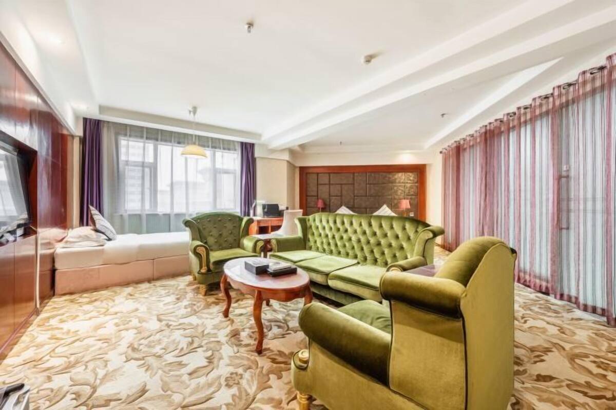 Habitación de hotel con un sofá verde, una mesita y un ventanal con cortinas.