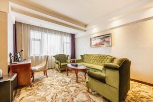 Habitación de hotel con sofá verde, dos sillones, un escritorio de madera y un ventanal con cortinas.