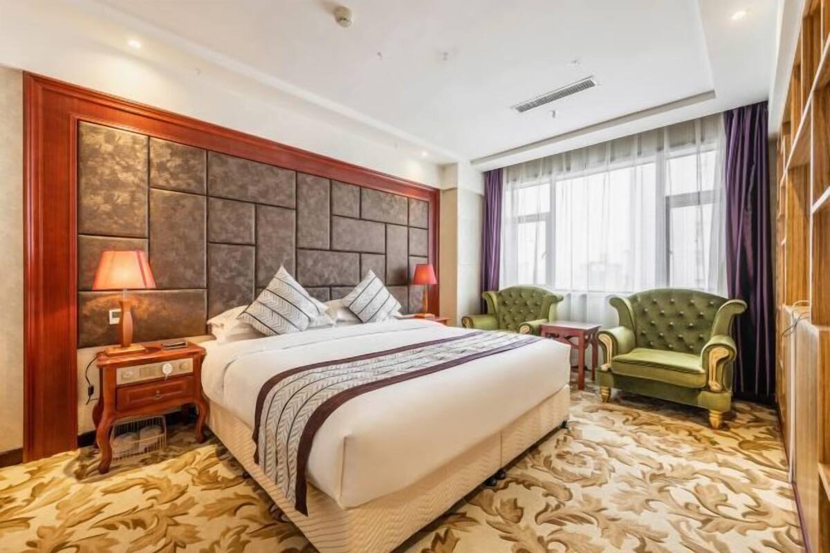 Habitación de hotel con una cama grande, dos mesitas de noche, un sillón verde y una ventana con cortinas.