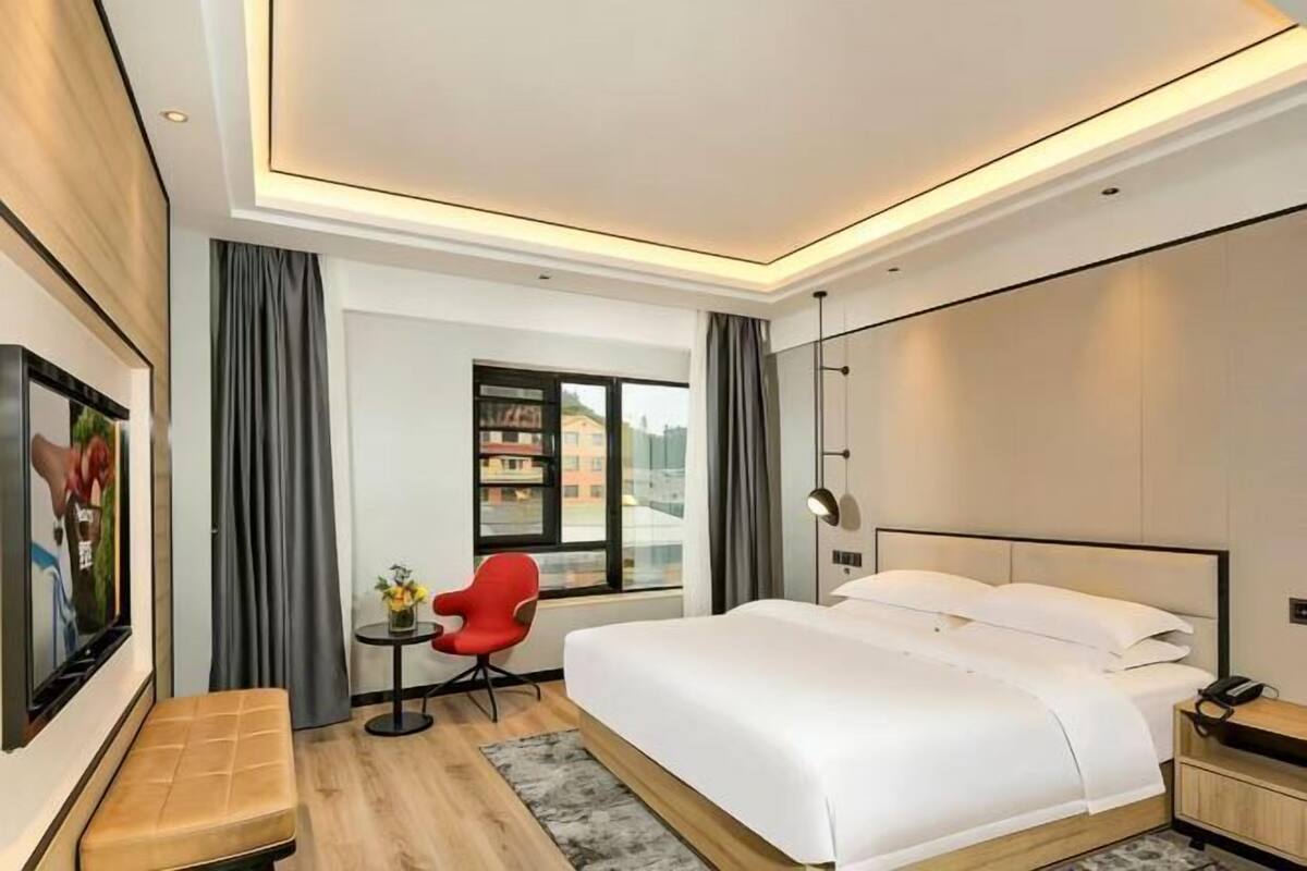 A modern hotel room with a large bed, a red chair, a small table, and a view through a window.
