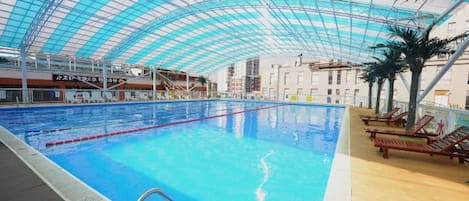 An indoor swimming pool with a glass ceiling, wooden deck, and lounge chairs.