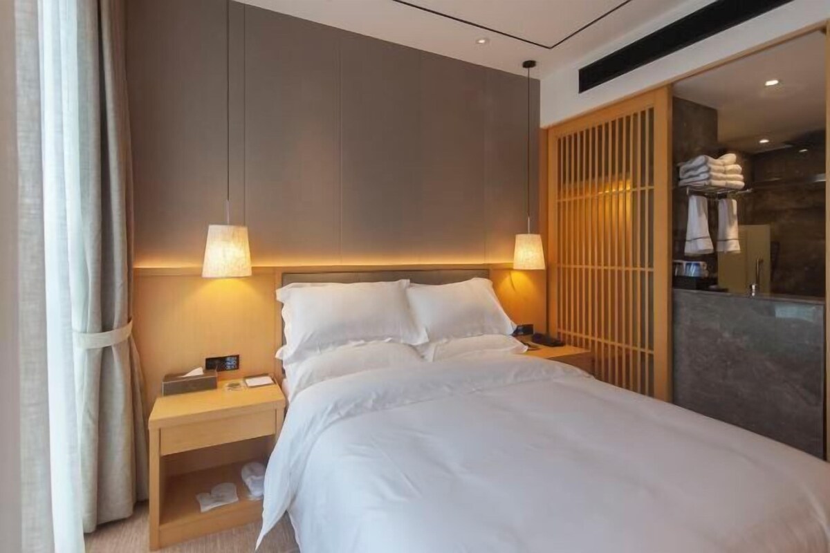 A hotel room with a bed, bedside lamps, a nightstand, a wooden partition, and a bathroom visible through a glass door.