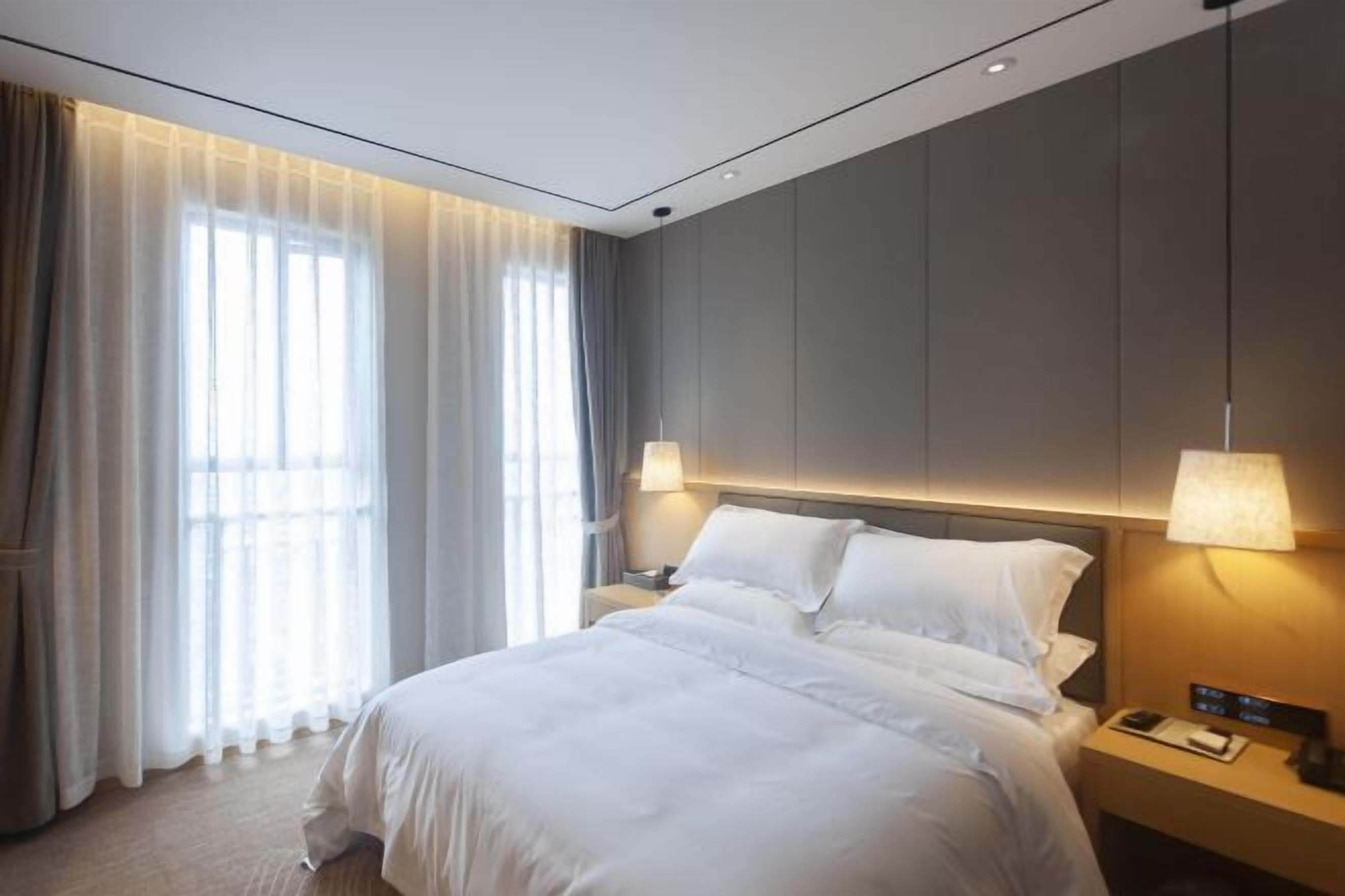 A modern hotel room with a large bed, two bedside lamps, a nightstand, and a window with sheer curtains.