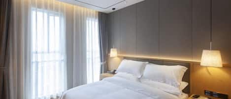 A modern hotel room with a large bed, two bedside lamps, a nightstand, and a window with sheer curtains.