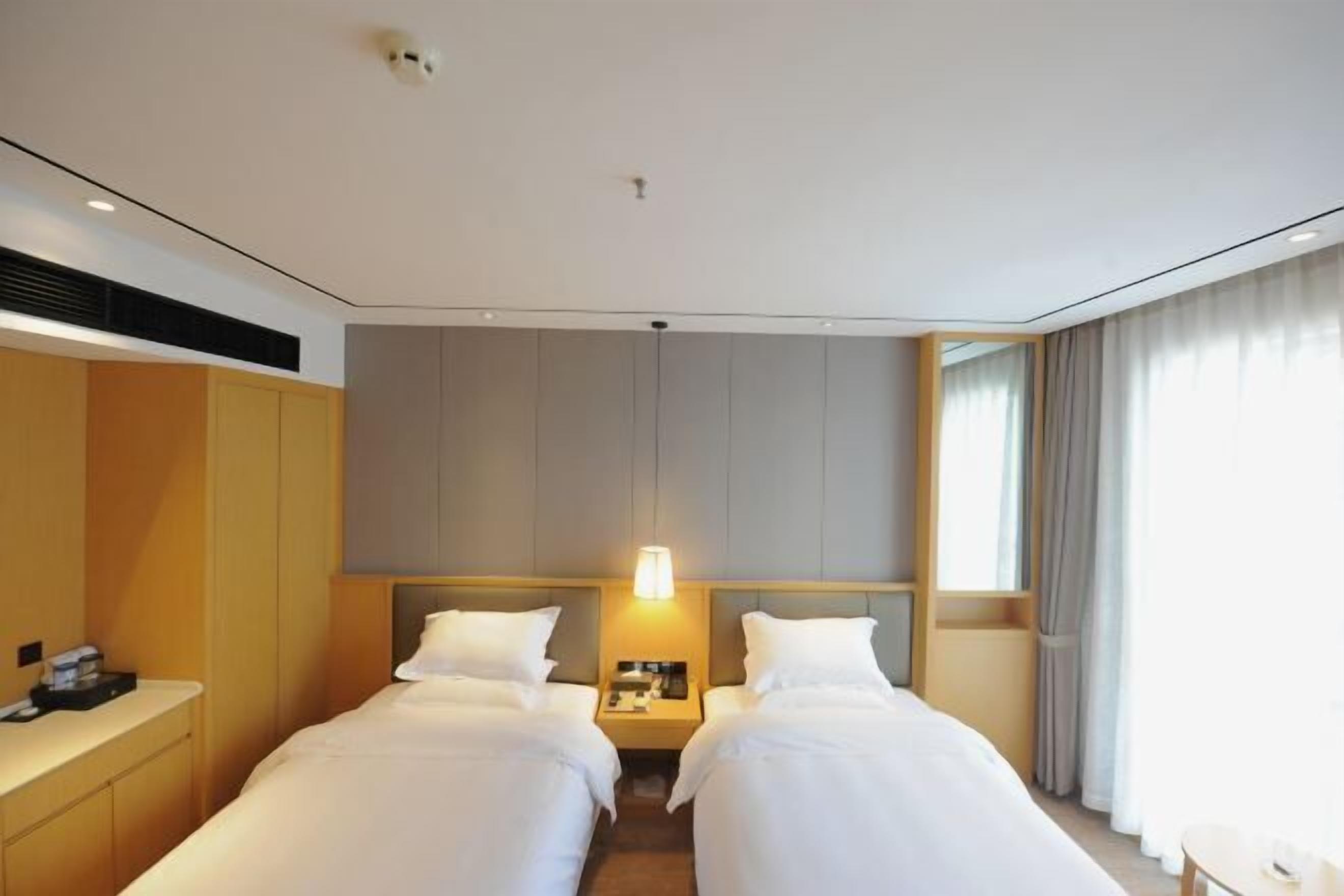 A hotel room with two beds, a bedside table with a lamp, a small desk, and a window with curtains.