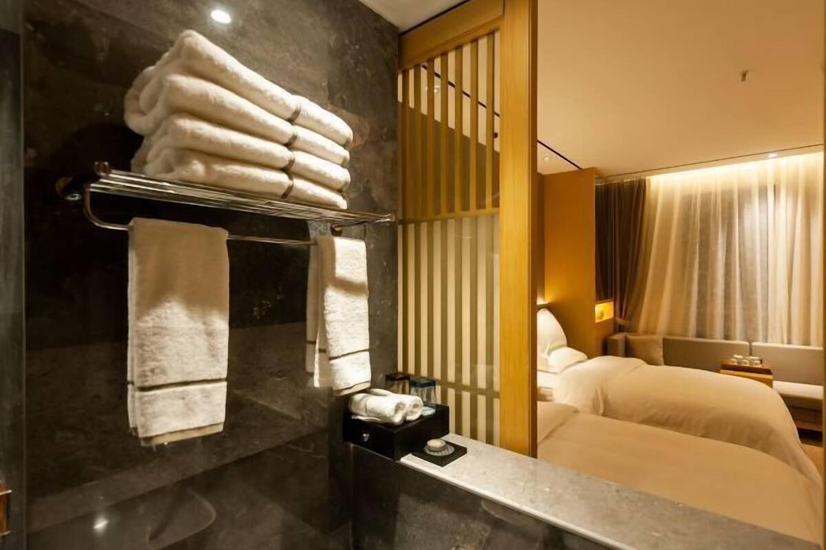 A hotel room with a marble wall, a towel rack with folded towels, a bedside table with a bottle, and a bed with a headboard.