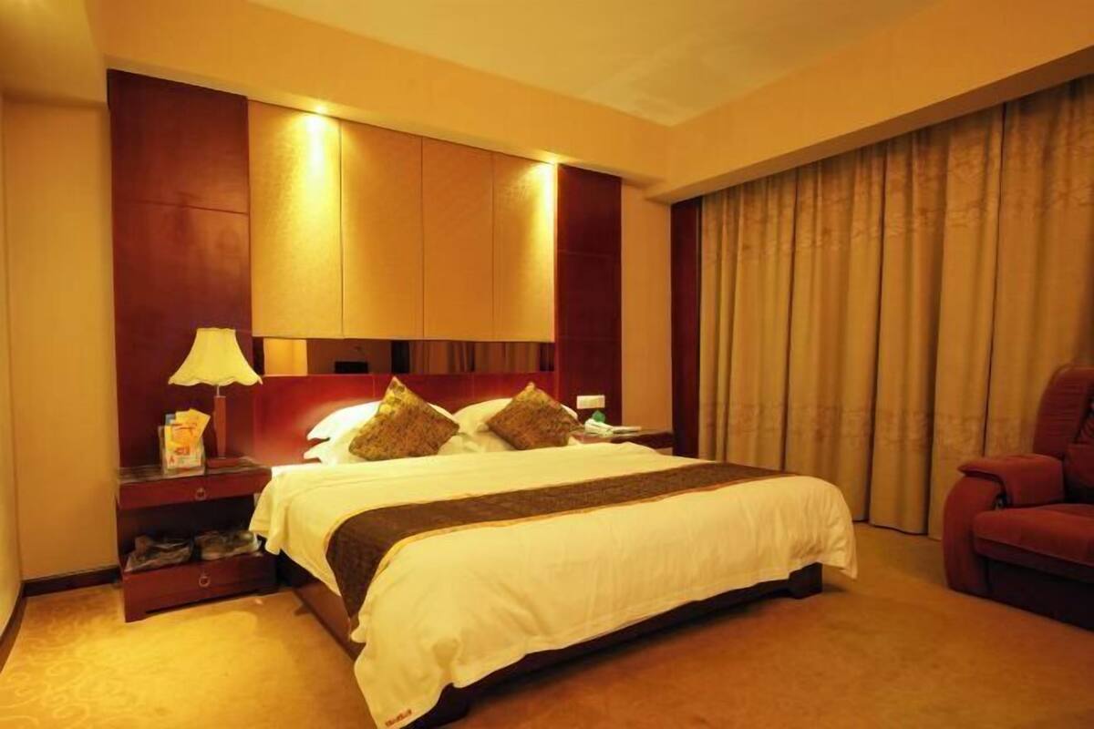 A hotel room with a large bed, a nightstand with a lamp, a red chair, and a large window with curtains.