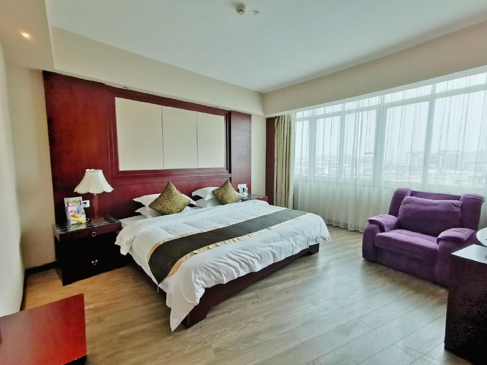 Een hotelkamer met een groot bed, een bureau met lamp, een paarse fauteuil en uitzicht door het raam.