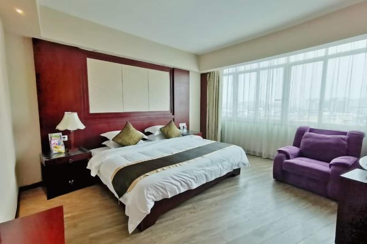 A hotel room with a large bed, a nightstand, a purple armchair, and a view of the city through large windows.