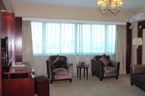 Une chambre d’hôtel avec une grande fenêtre, deux fauteuils, une petite table et un bureau avec une chaise.