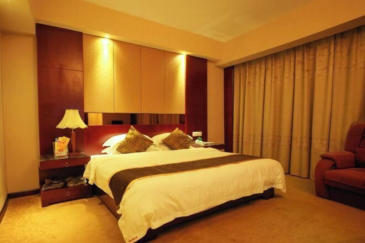Une chambre d’hôtel avec un grand lit, une table de chevet avec une lampe, un fauteuil rouge et une fenêtre avec des rideaux.