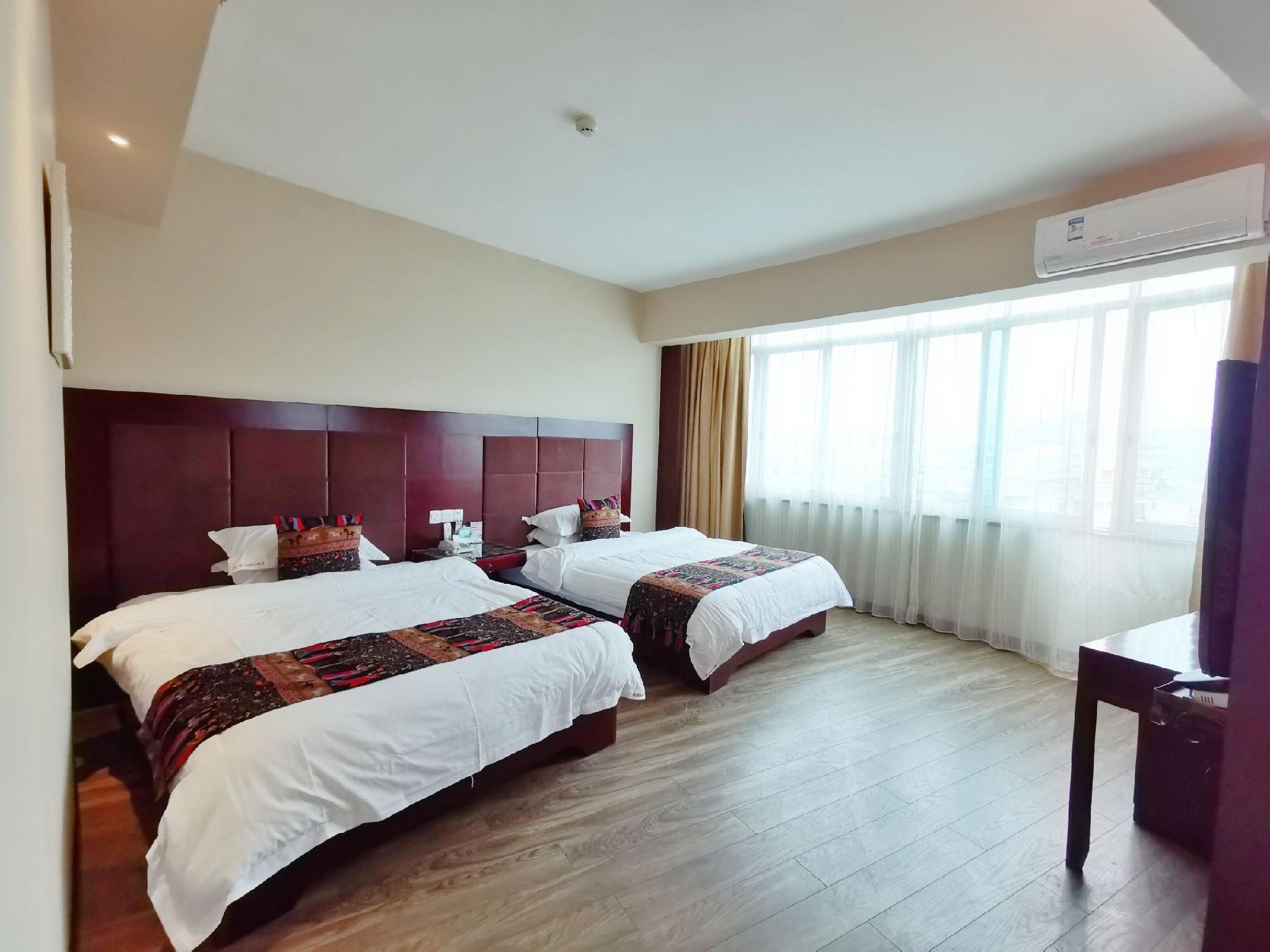 A hotel room with two beds, a wooden floor, a large window with curtains, and an air conditioning unit.