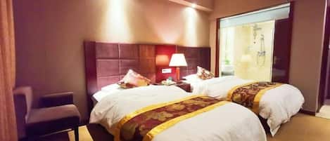 A hotel room with two beds, a chair, a nightstand with a lamp, and a bathroom visible through a glass door.