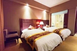 Habitación de hotel con dos camas, una silla, una mesita de noche con lámpara y un baño visible a través de una puerta de cristal.