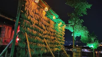 A nighttime outdoor area with a structure covered in foliage, illuminated by warm and green lights, and a paved walkway with benches.