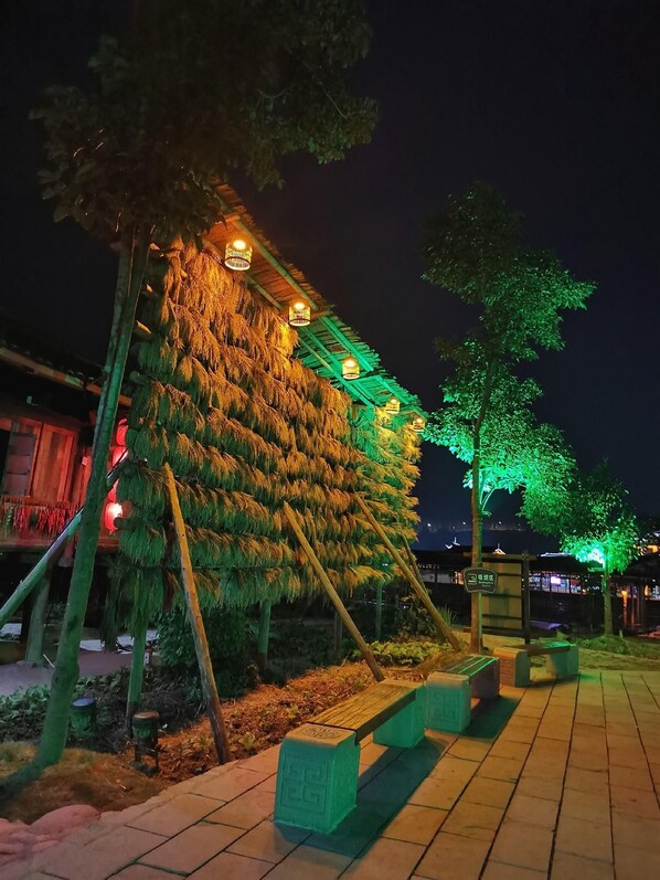 Un espacio exterior nocturno con una estructura cubierta de follaje, iluminada con luces cálidas y verdes, y un sendero pavimentado con bancos.