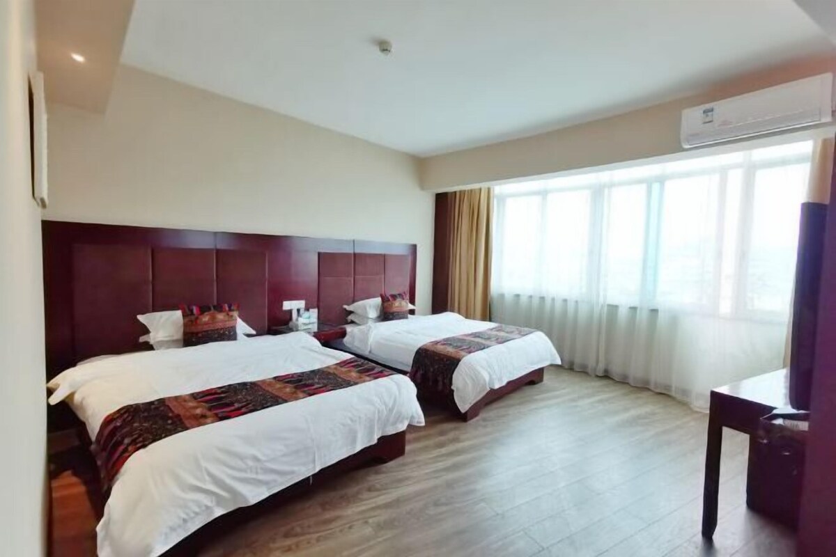 A hotel room with two beds, a wooden floor, a large window with curtains, and an air conditioning unit.