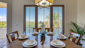 Un coin repas avec une grande table dressée pour six personnes, de grandes fenêtres équipées de stores et un luminaire suspendu.