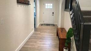 Un passadís amb sòl de fusta, una banca, una planta en test i una porta, i una escala amb barana negra.