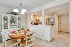 A dining area with a wooden table and white chairs, a white cabinet with glass doors, and a kitchen area with white cabinets and stainless steel appliances.