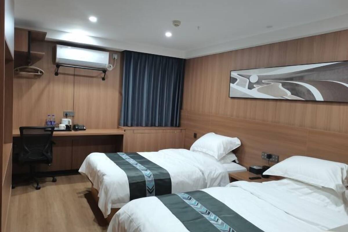 Habitación de hotel con dos camas, un escritorio, una silla, un equipo de aire acondicionado y un cuadro en la pared.