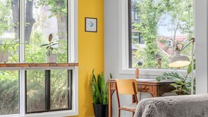 Una habitación con una pared amarilla, un escritorio de madera, una silla, una lámpara y vistas a árboles y edificios.