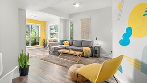 Una sala de estar moderna con un sofá gris, un sillón amarillo y una mesita. Se ve una ventana grande con vistas a espacios verdes.