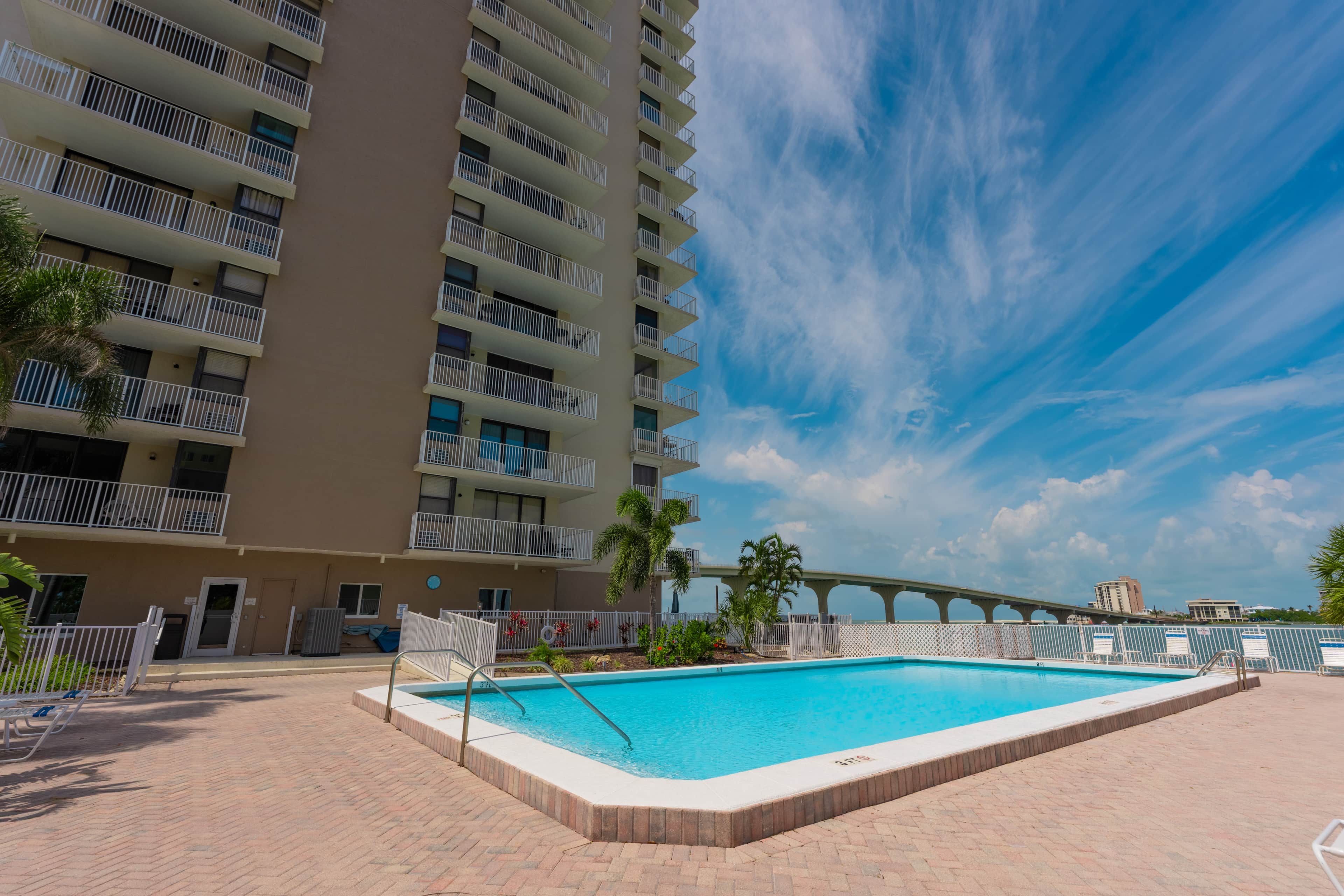 A swimming pool with a clear blue sky in the background, surrounded by a brick patio and a multi-story building with balconies.