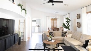 A living room with a sofa, coffee table, and TV. There is a ceiling fan, plants, and a dining area in the background. - The Gilbert Haven - comfortable, stylish w/ pool (Gilbert)