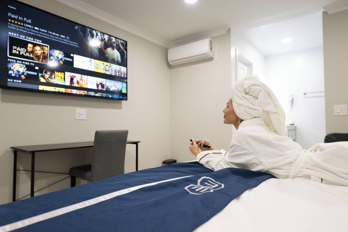 A hotel room with a bed, a TV mounted on the wall displaying various channels, a desk with a chair, and an air conditioning unit.