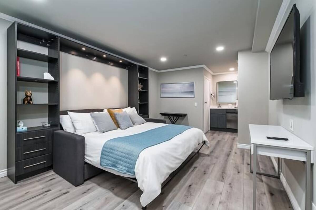 A modern bedroom with a large bed, built-in shelving, a small desk, and a TV.