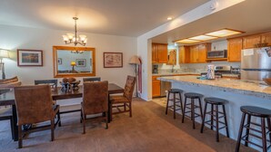A modern kitchen with wooden cabinets, a central island with stools, a dining area with chairs, and a chandelier. - Interlaken #19 - 3 Bedroom 3 Bathroom Unit (June Lake)