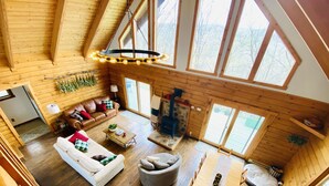 A spacious loft-style room with a high ceiling, wooden walls, and large windows. There is a cozy seating area with a sofa and armchairs, a fireplace, and a dining table.
