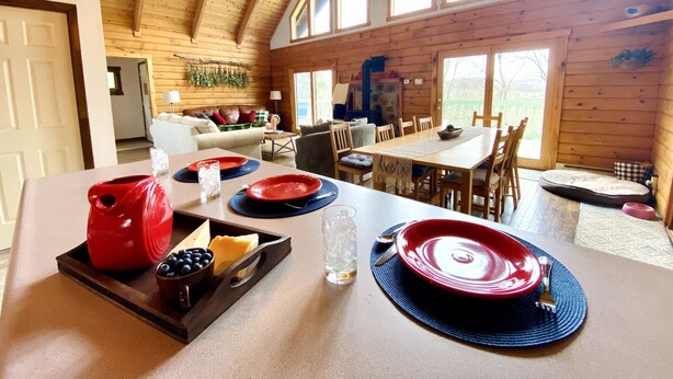 A wooden cabin interior with a dining area set for four, featuring a table with red and blue plates, a bowl of blueberries, and a cheese platter.