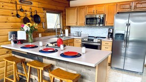 A modern kitchen with wooden cabinets, a stainless steel refrigerator, a microwave, and a stove. There are bar stools and a large island with plates and glasses.