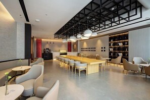 Un hall d’hôtel moderne avec une longue table commune, des espaces aménagés pour s’asseoir et un coin bar.