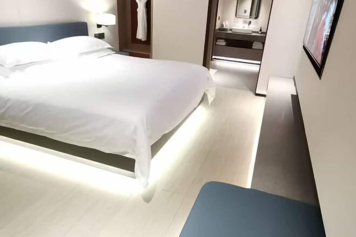 Moderni hotellihuone, jossa on sänky, pieni oleskelualue, kylpyhuone, jossa on pesuallas ja peili, sekä seinällä oleva kehystetty taulu.