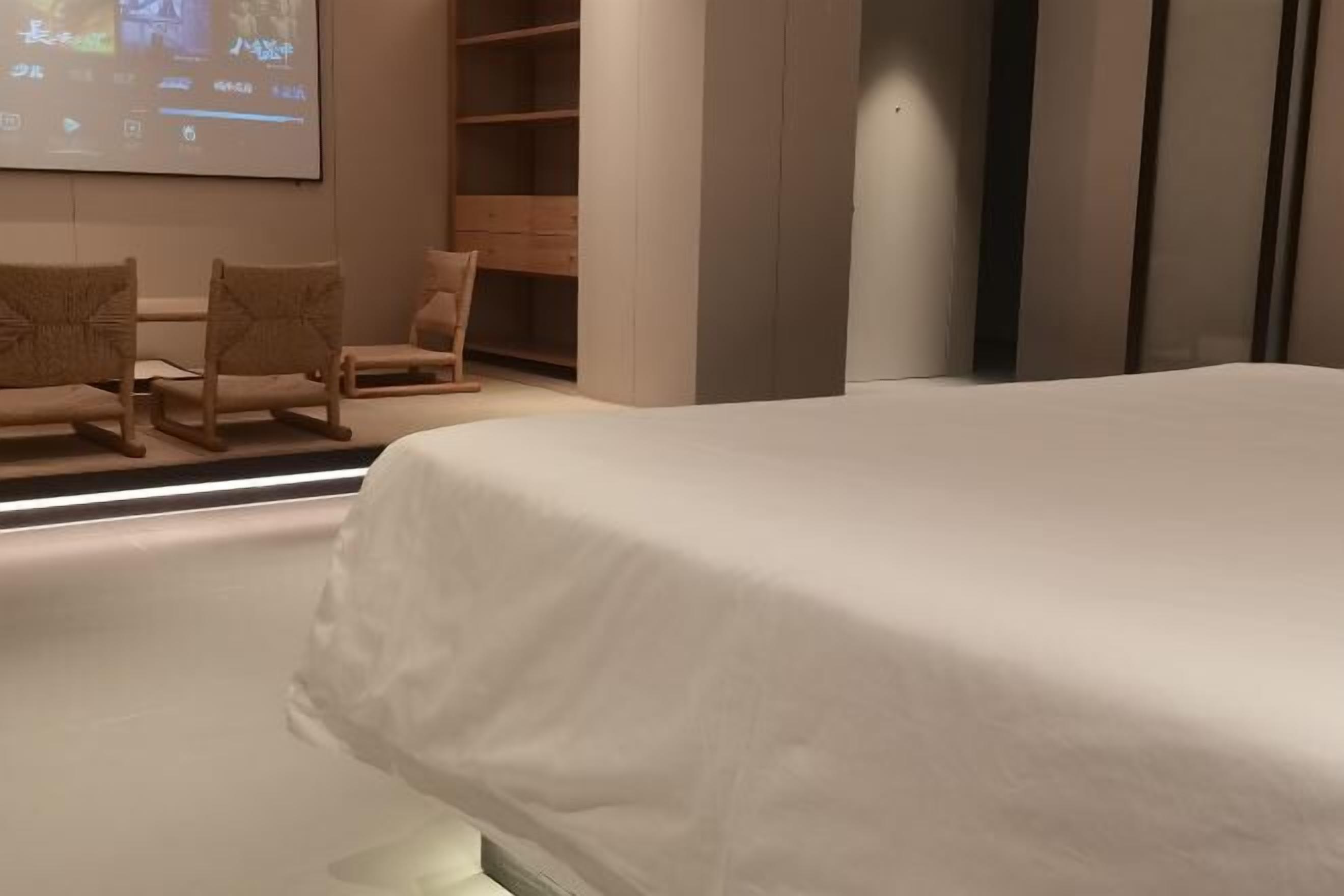 Habitación de hotel con cama, zona de estar con sillones, armario de madera y una pantalla montada en la pared.