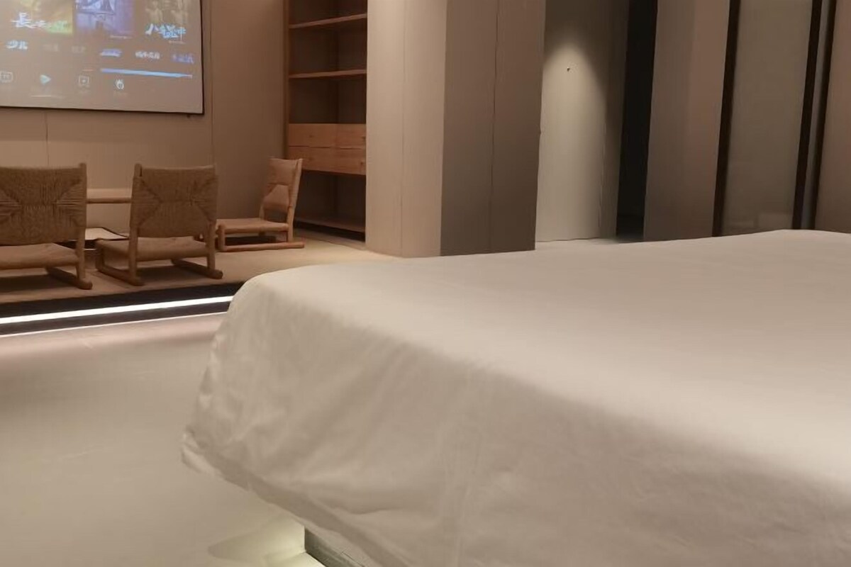 A hotel room with a bed, a seating area with chairs, a wooden wardrobe, and a wall-mounted screen.