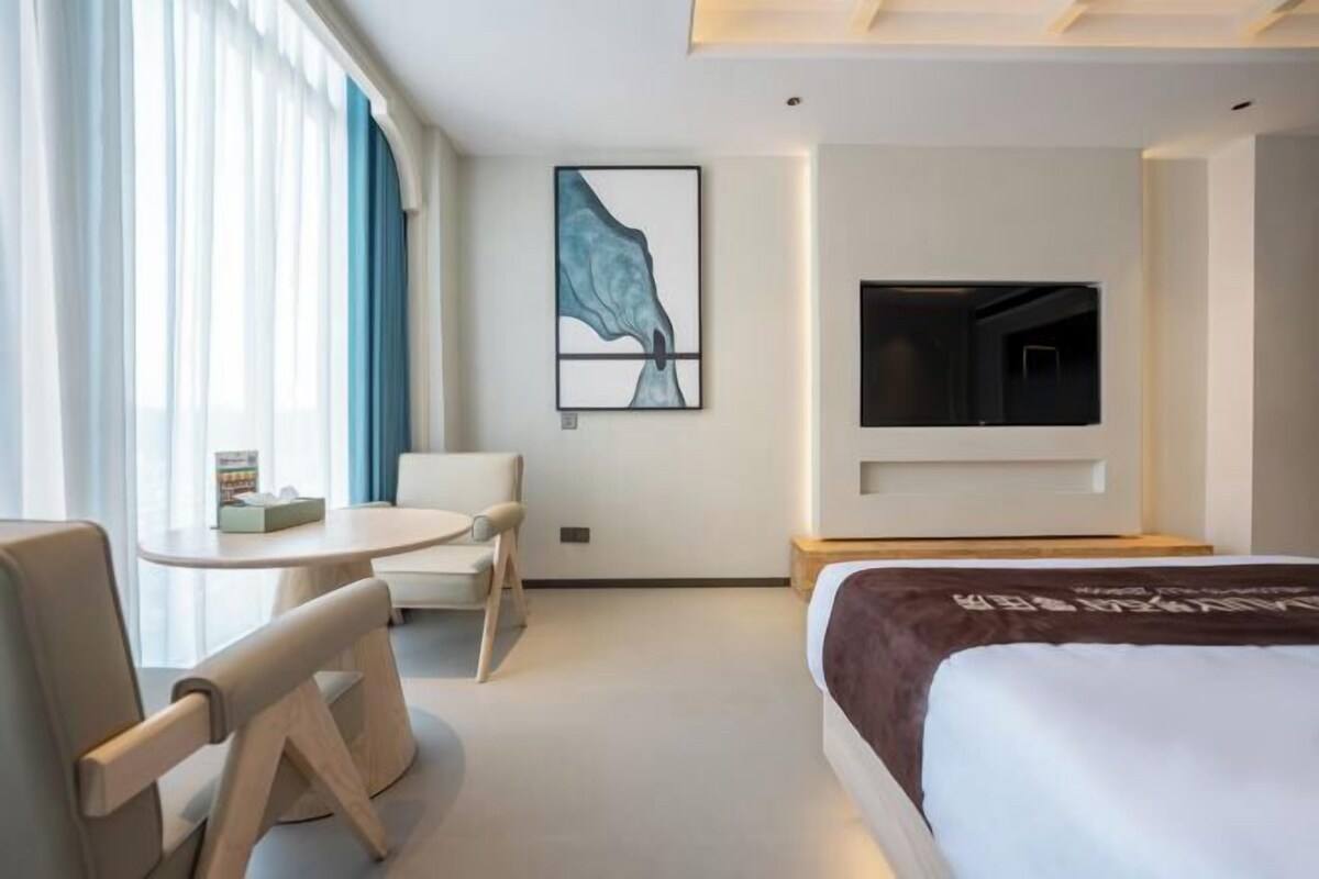 A modern hotel room with a bed, a round table, a chair, a TV, and a painting on the wall.