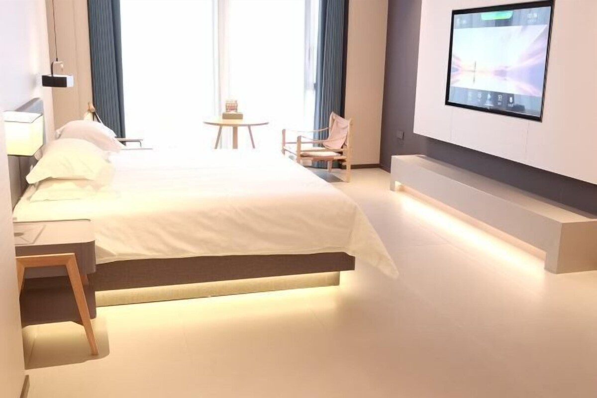 A modern hotel room with a bed, bedside table, chair, TV, and a small table with a lamp.